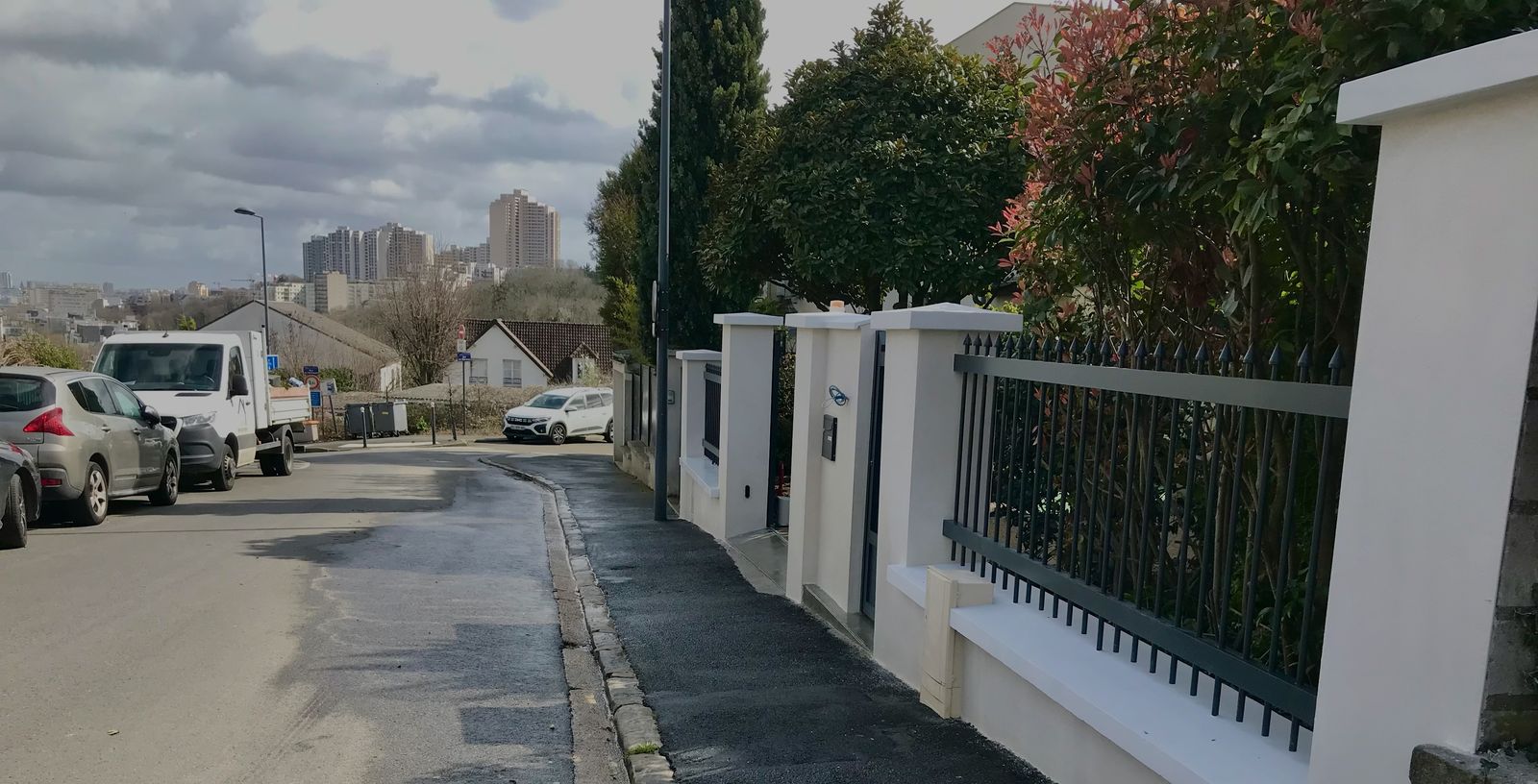 Grille en aluminium noir sur muret blanc, le long d'une rue en pente